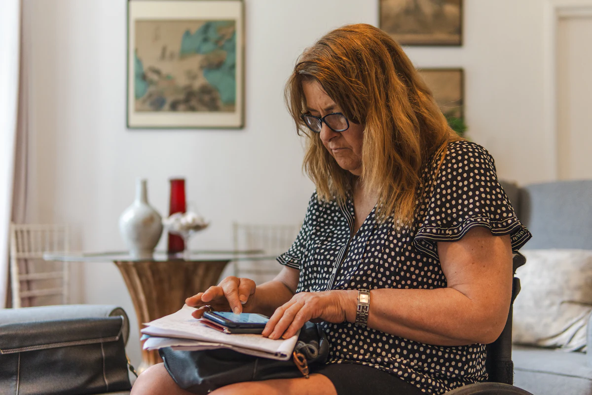 A woman at home writing figures on paper while checking her phone.