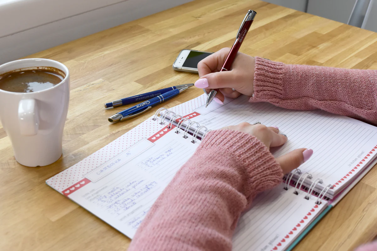 A person writing in a notebook at a wooden table with a cup of coffee.