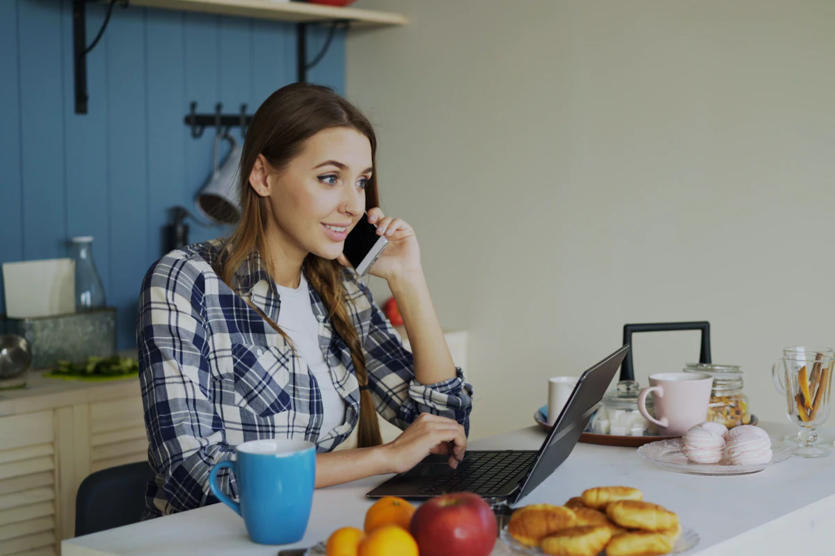 A woman on the phone while using a laptop at her kitchen table.