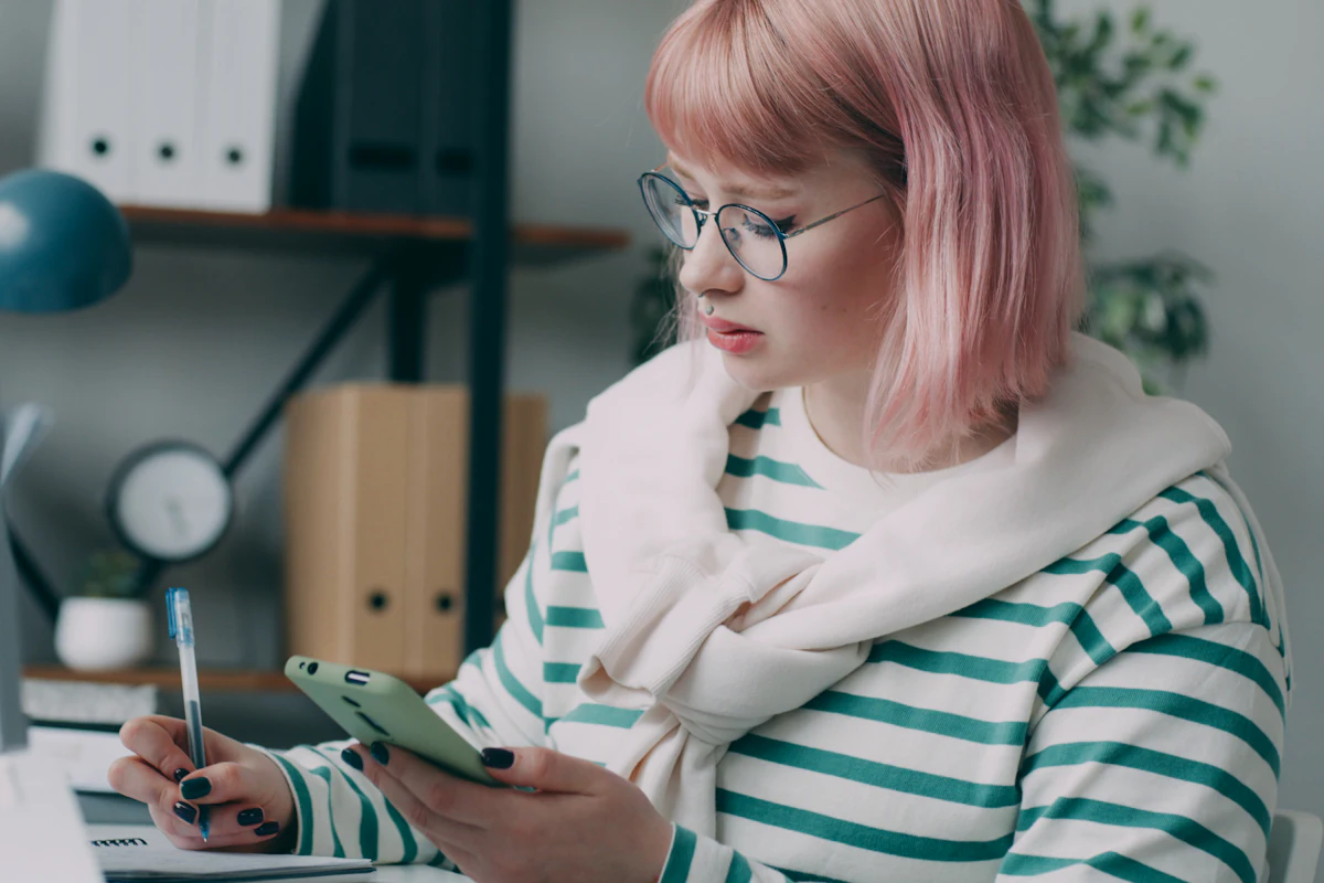 A woman with glasses writing notes while checking her phone at a desk.
