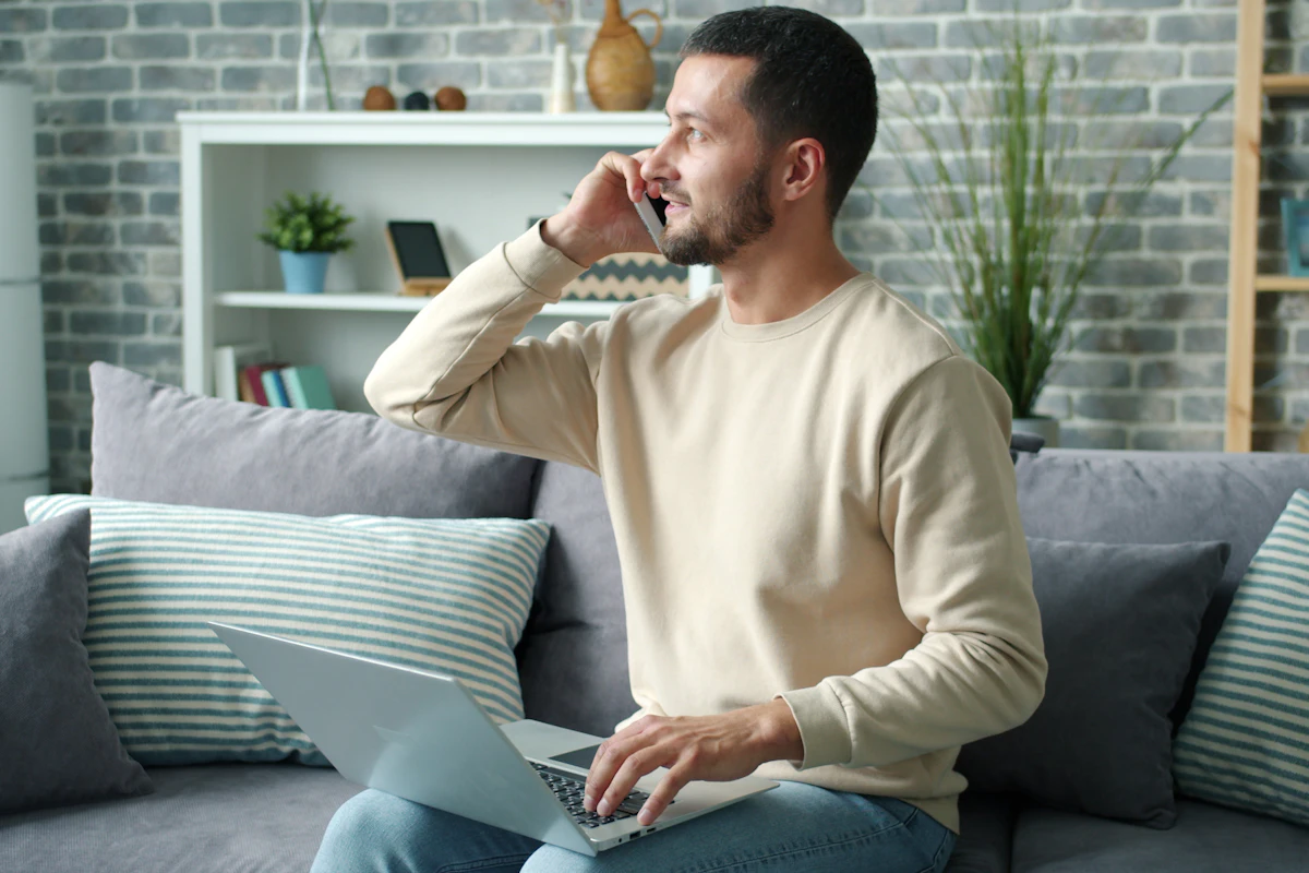 A man on the phone with a laptop on his lap, sitting on a sofa.